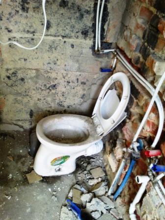 Stary, brudny sedes z otwartą klapą w trakcie demontażu w pomieszczeniu z odsłoniętą cegłą i gruzem na podłodze. Widoczne rury instalacji wodnej.