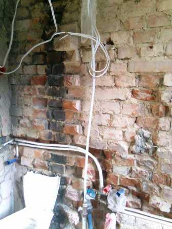 Niefotogeniczna ściana z czerwonej cegły z nowymi rurami i przewodami elektrycznymi w trakcie prac instalacyjnych.