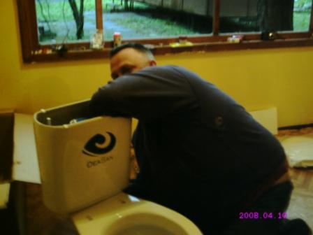 Mężczyzna w trakcie montażu spłuczki toalety marki Deaksan w łazience o żółtych ścianach, widoczne okno i data wykonania zdjęcia.