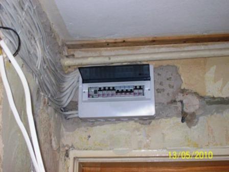 Otwarta skrzynka elektryczna z licznymi kablami na ścianie w trakcie remontu, widoczne ślady po tapetach i niedokończone prace wykończeniowe.