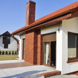 Elewacja domu z ceglanym kominem, brązowymi oknami i częściowo opuszczoną roletą zewnętrzną w kolorze brązu, widok z tarasu.