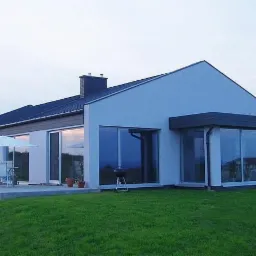 Nowoczesny dom jednorodzinny z dużymi oknami PCV i drzwiami przesuwnymi, widok z zewnątrz na trawnik i patio z parasolem.