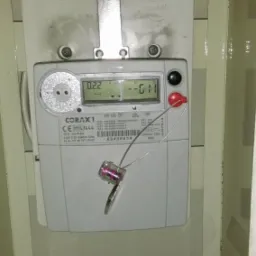 Zamknięty licznik elektryczny Corax 1 w metalowej skrzynce z plombą zabezpieczającą, widoczny numer seryjny i wskazania wyświetlacza.