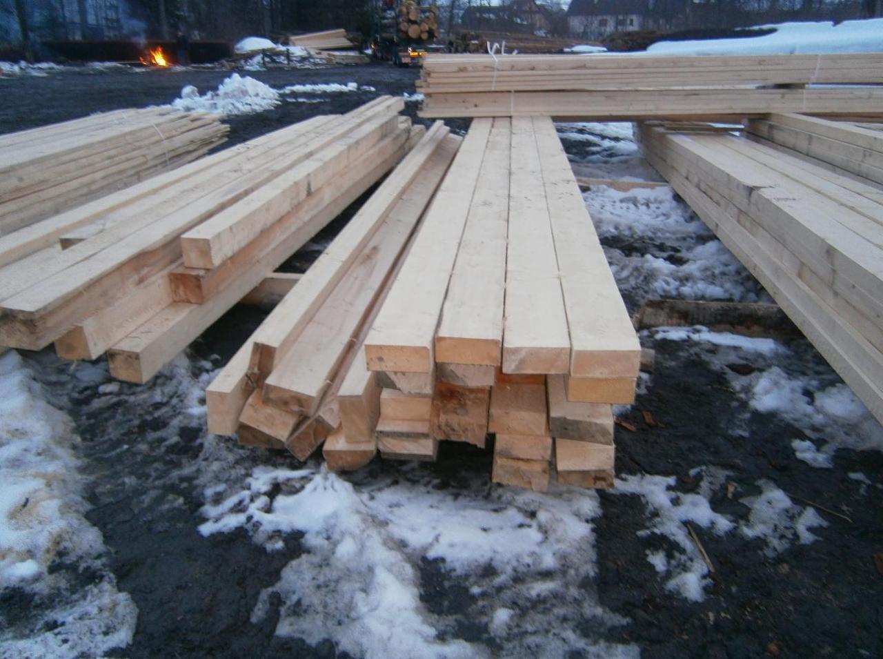 Stosy świeżo przetartego drewna budowlanego leżące na ziemi z resztkami śniegu, w tle ognisko i budynki.