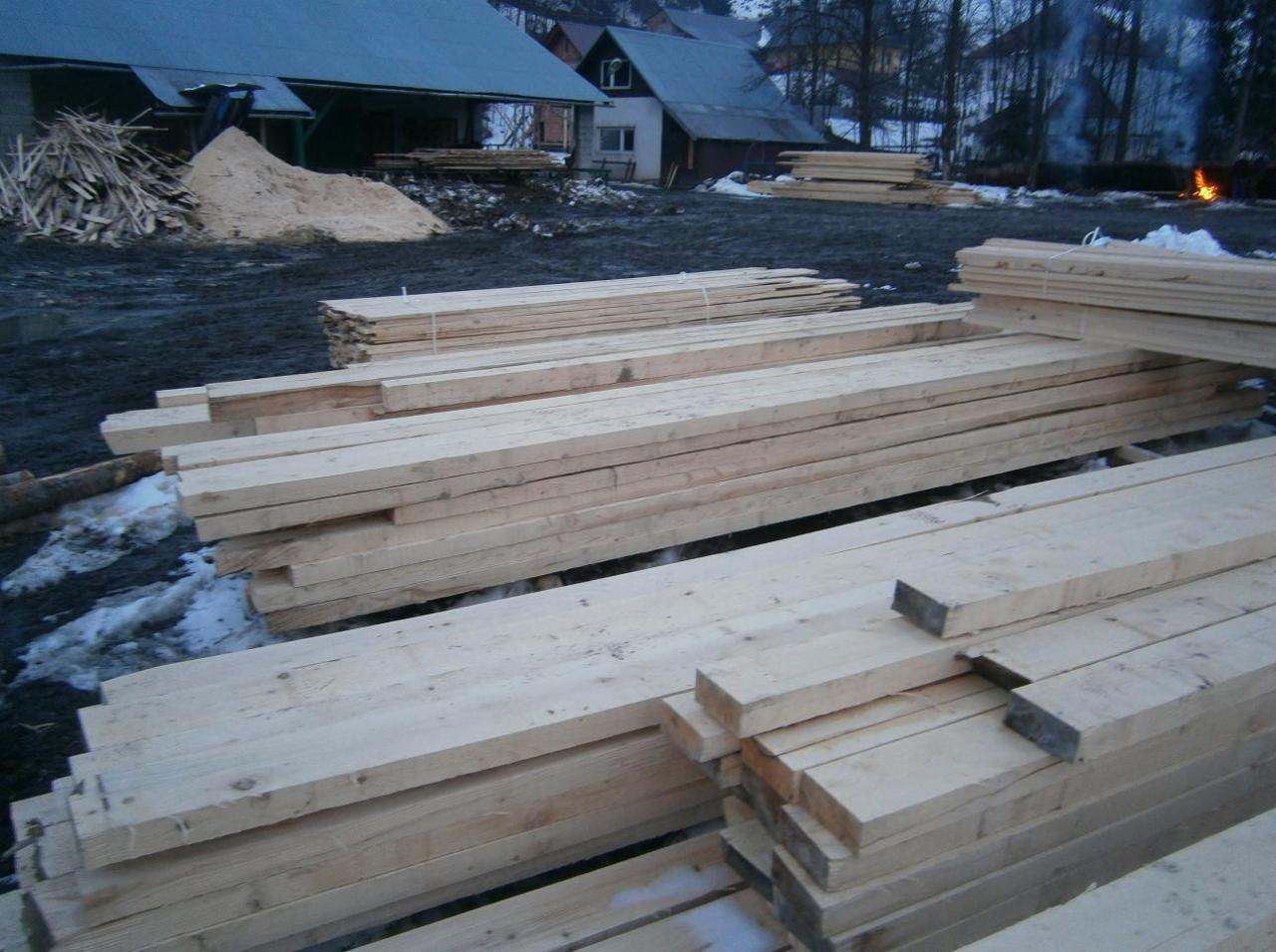 Stosy świeżo przetartego drewna budowlanego na placu składowym, w tle budynki mieszkalne, sterta trocin i palące się ognisko.