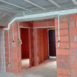 Wnętrze w trakcie budowy z widocznymi przewodami elektrycznymi ułożonymi na ścianach z czerwonej cegły i pod betonowymi schodami.