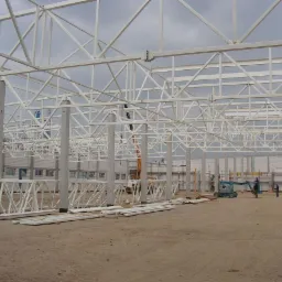 Wnętrze budowanej hali z widoczną białą konstrukcją szkieletową, betonowymi słupami i elementami ścian z płyt warstwowych. Na ziemi leżą przygotowane profile. W tle widoczny podnośnik koszowy...