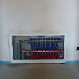 Rozdzielacz ogrzewania podłogowego w skrzynce, z czerwonymi i niebieskimi rurami, widoczne zawory i oznaczenia 'Powrót' i 'Zasilanie', zamontowany na tle surowej ściany i betonowej podłogi.