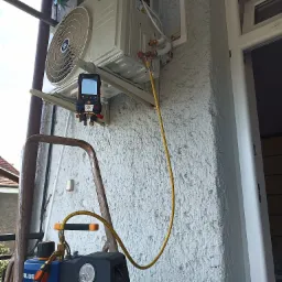 Zewnętrzna jednostka pompy ciepła Kaisai GEO podłączona do pompy próżniowej VALUE za pomocą żółtego węża, manometry cyfrowe Testo 557s zawieszone na jednostce, ściana z grubym tynkiem, drabina...