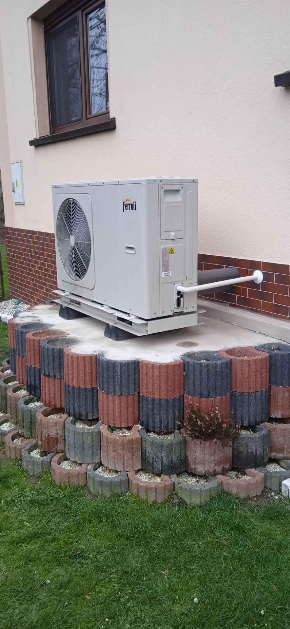 Zewnętrzna jednostka pompy ciepła Ferroli, umieszczona na betonowym fundamencie obłożonym dwukolorowymi donicami z roślinami, przy ścianie budynku z oknem.