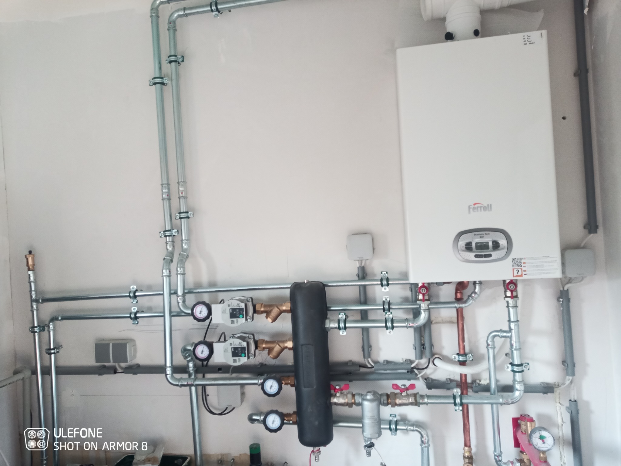 Biały piec gazowy Ferroli z podłączonymi rurami miedzianymi i stalowymi, manometry na instalacji, zbiornik wyrównawczy pionowy, całość na tle białej ściany.