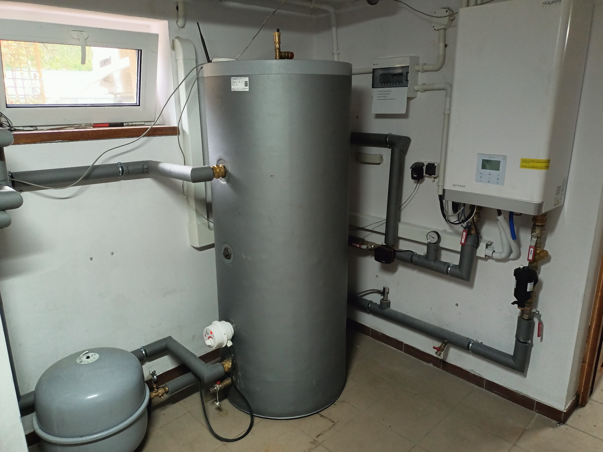 Szary zbiornik akumulacyjny ciepła z podłączonymi szarymi rurami, obok biały piec gazowy Aquami Rapid i licznik elektryczny w pomieszczeniu z oknem.