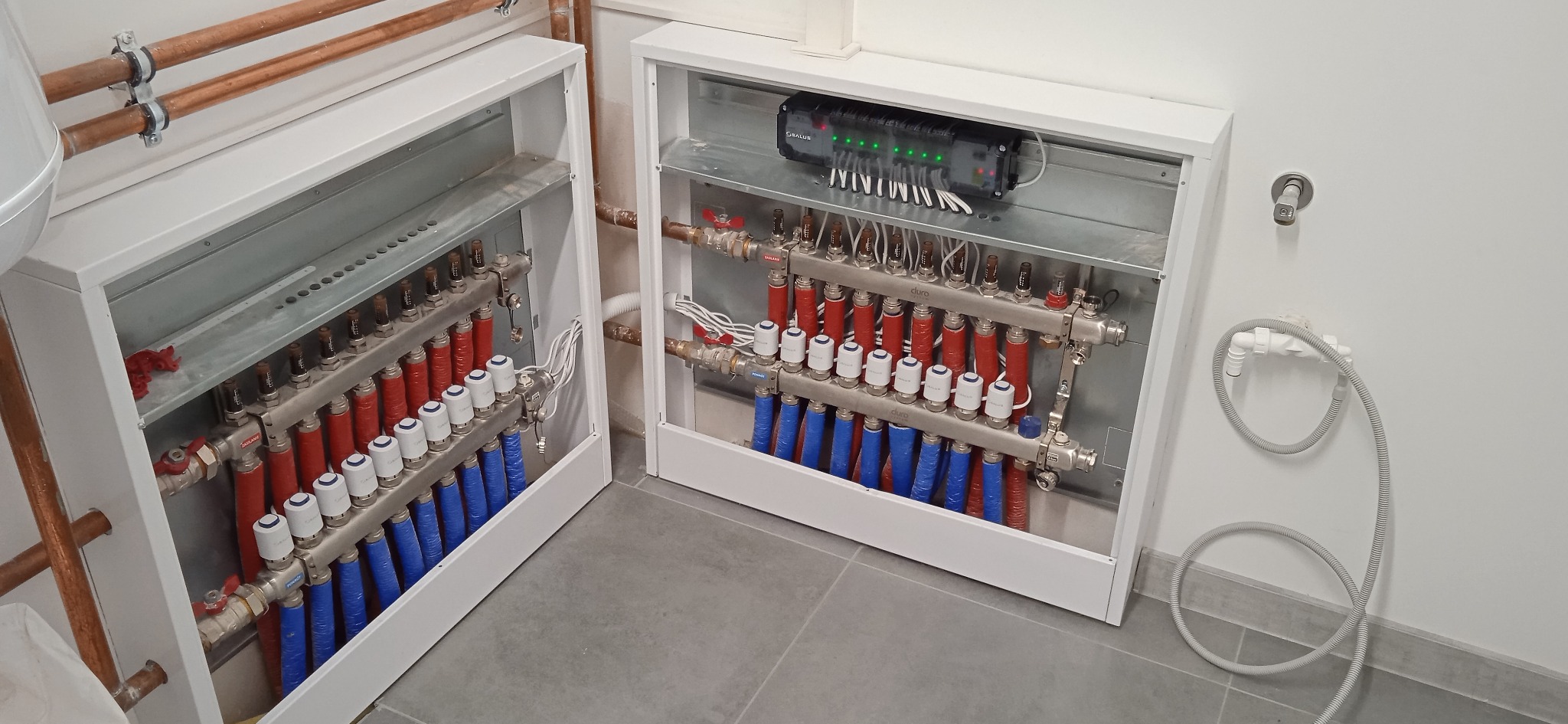 Dwa rozdzielacze ogrzewania podłogowego w białych obudowach, z rurami w kolorach czerwonym i niebieskim, z widocznym sterownikiem elektronicznym z zielonymi diodami, zamontowane na tle szarej...