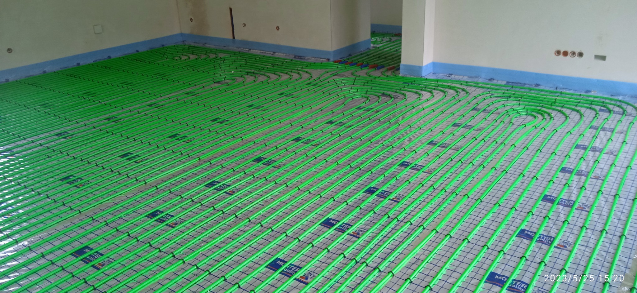 Zielone rury ogrzewania podłogowego ułożone na siatce w surowym pomieszczeniu z niepomalowanymi ścianami i niebieskim paskiem ochronnym przy podłodze.