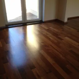 Błyszcząca, świeżo cyklinowana parkietowa podłoga w odcieniach brązu, z widocznym odbiciem światła słonecznego wpadającego przez okno balkonowe.