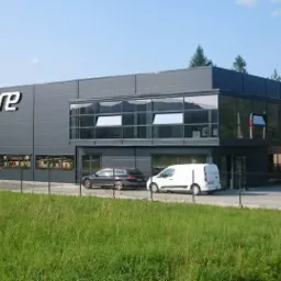 Nowoczesny budynek usługowy z szarej blachy trapezowej, duże okna z refleksami, logo firmy na elewacji, samochody zaparkowane przed budynkiem, otoczenie: trawa i drzewa.