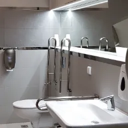Toaleta publiczna przystosowana dla osób niepełnosprawnych, z umywalką, uchwytami i dozownikiem mydła, wykończona w jasnych szarościach z heksagonalną fakturą płytek.
