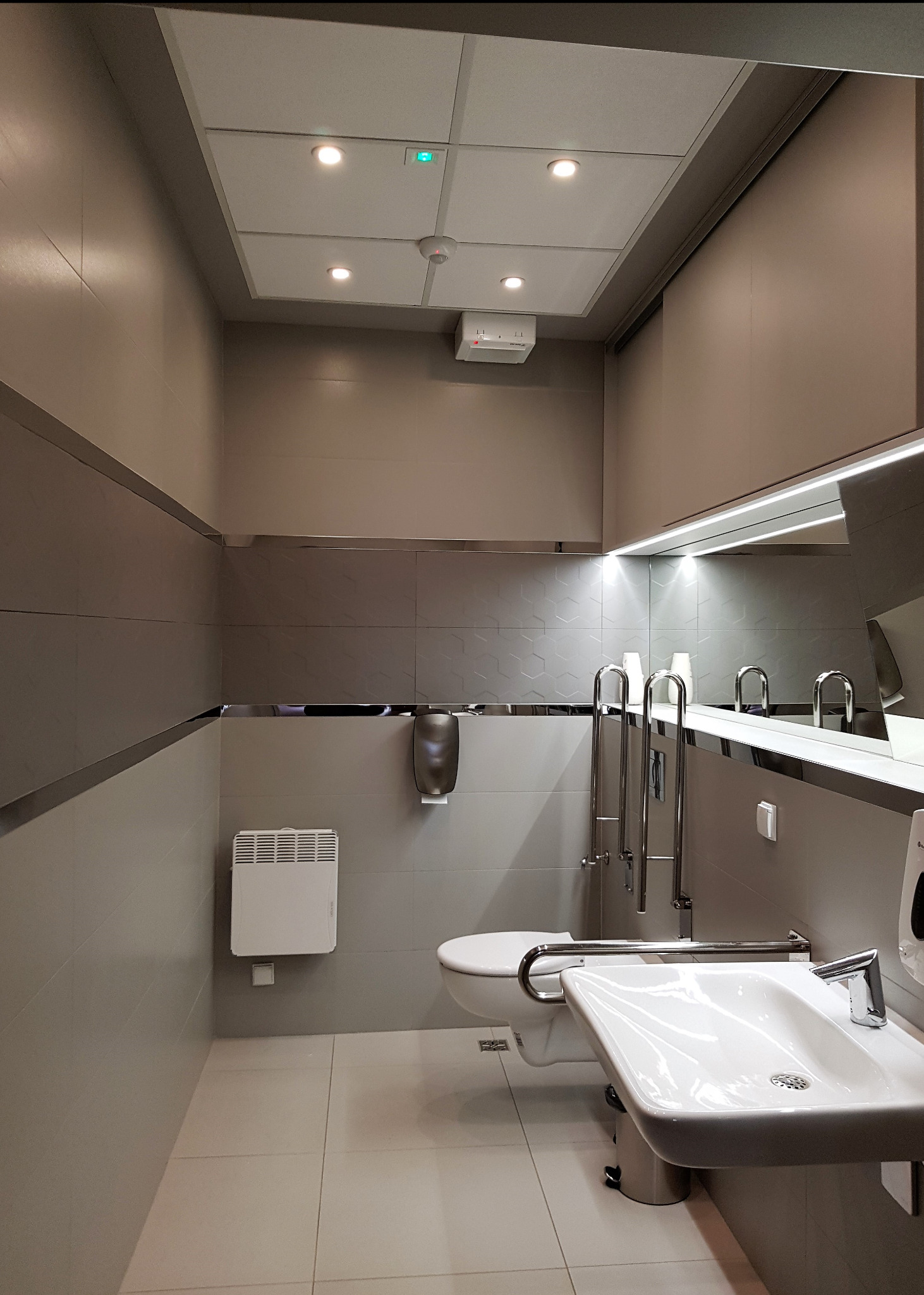 Nowoczesna łazienka publiczna z udogodnieniami dla niepełnosprawnych: umywalka, toaleta z poręczami, podajnik ręczników i grzejnik elektryczny, wykończona w szarościach z akcentami chromu.