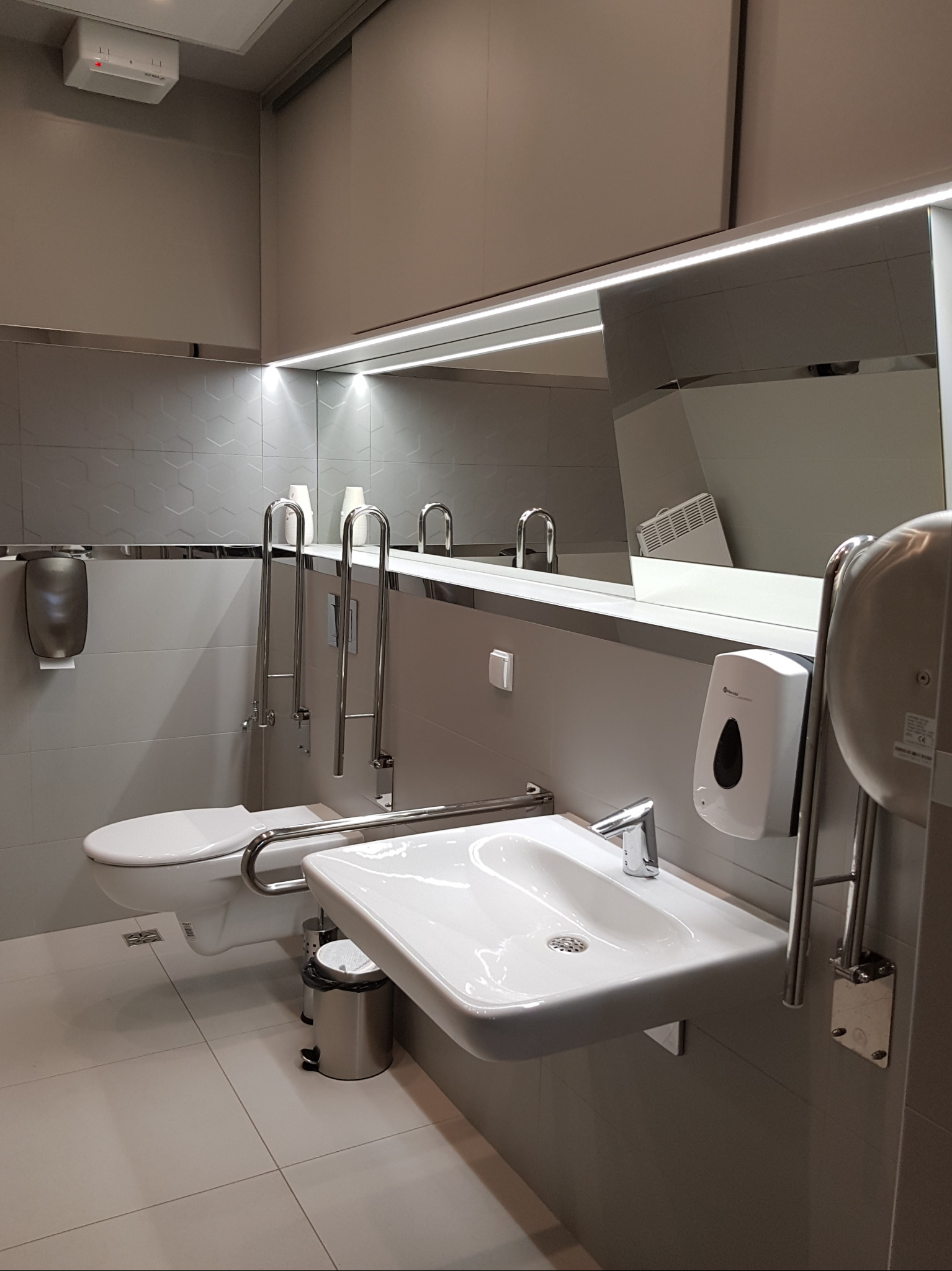 Toaleta publiczna przystosowana dla osób niepełnosprawnych, z umywalką, toaletą i uchwytami ze stali nierdzewnej, odbicie grzejnika w lustrze.