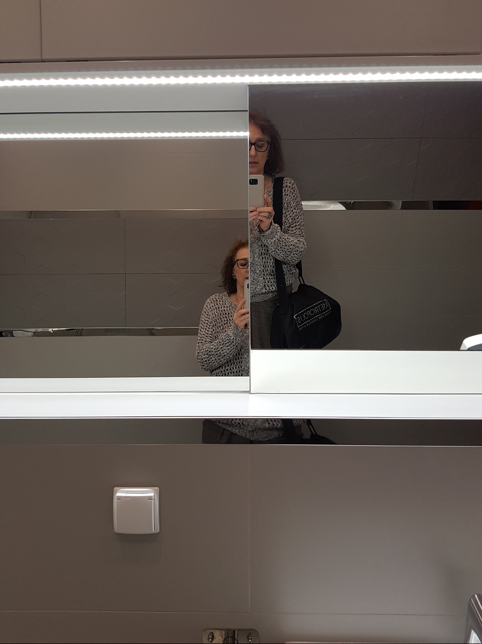 Kobieta w okularach robi selfie w łazience z podświetlanym lustrem i szarymi płytkami.