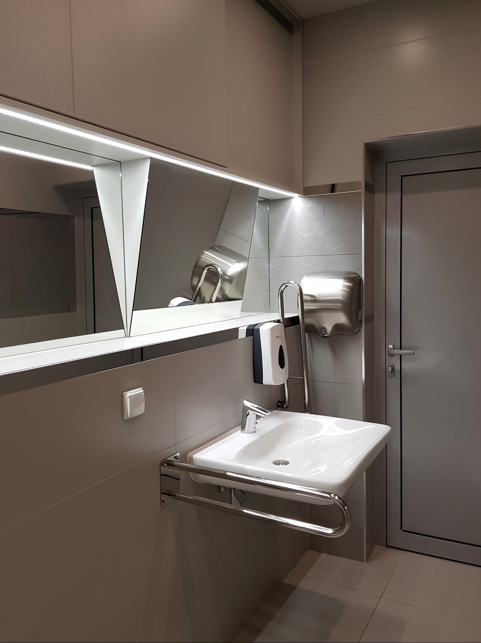 Nowoczesna umywalka z chromowanymi uchwytami dla niepełnosprawnych, biała ceramika, srebrny kran, dozownik mydła i suszarka do rąk na tle heksagonalnych płytek w odcieniach szarości.