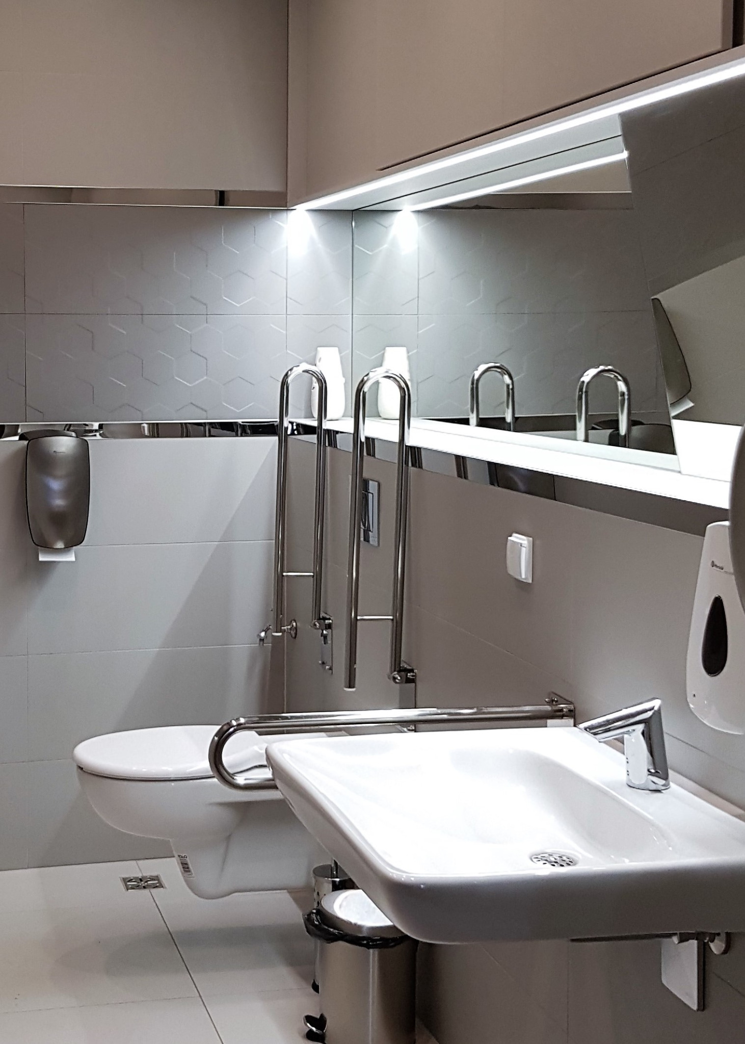 Toaleta publiczna przystosowana dla osób niepełnosprawnych, z umywalką, uchwytami i dozownikiem mydła, wykończona w jasnych szarościach z heksagonalną fakturą płytek.