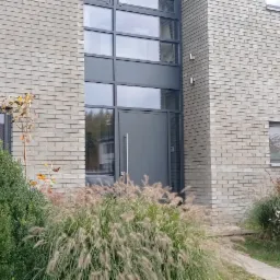 Szary, nowoczesny dom z cegły z dużymi oknami i szarą stolarką, otoczony ozdobnymi trawami i krzewami.