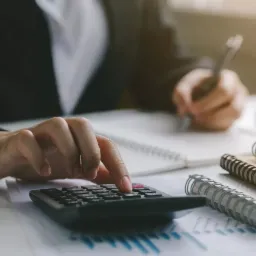 Kobieta w czarnym żakiecie kalkuluje coś na kalkulatorze, jednocześnie notując w notesie. Na biurku leżą dodatkowe notesy i dokumenty z wykresami.