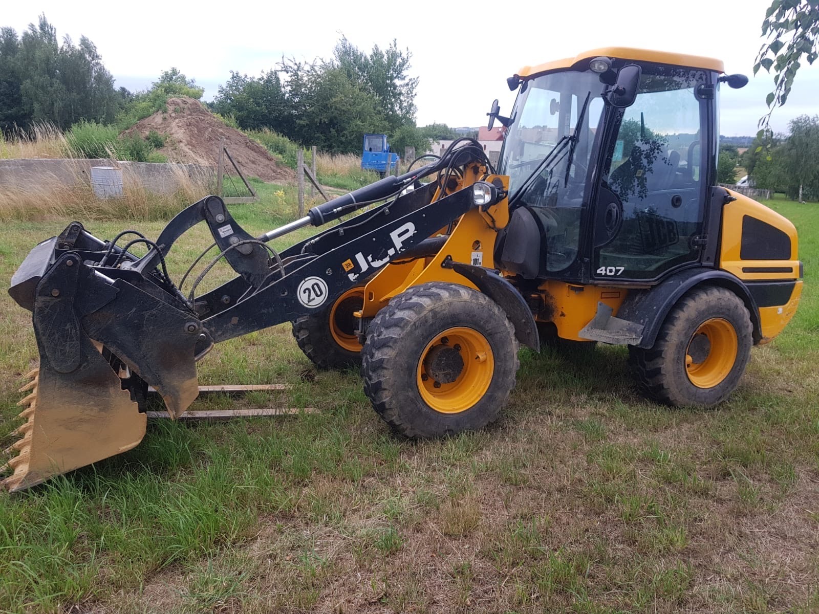 Żółto-czarna ładowarka JCB 407 na trawiastym terenie, z widocznym wykopem ziemi w tle. Sprzęt gotowy do pracy, z detalami jak opony i kabina w ostrym świetle.
