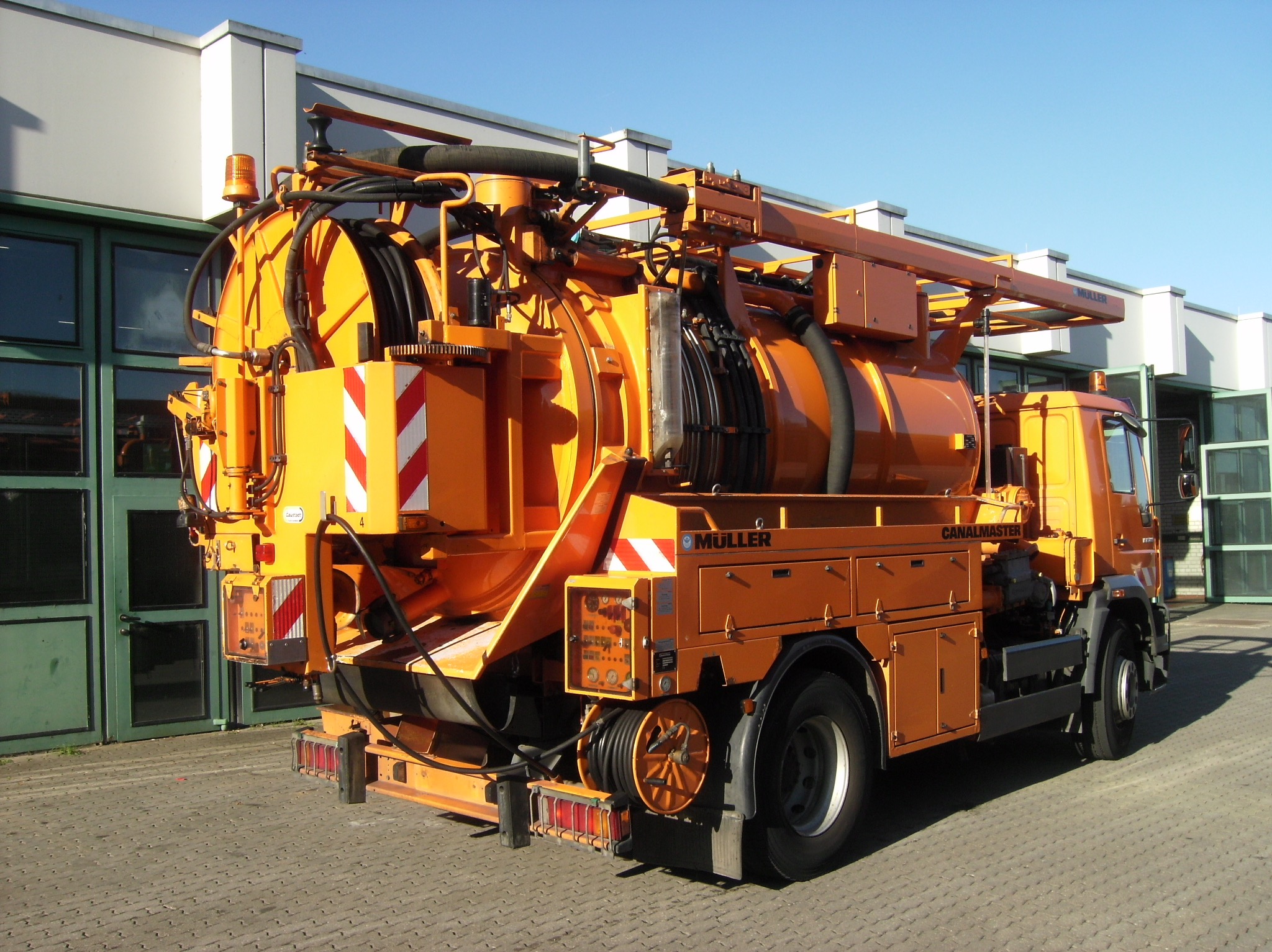 Pomarańczowy samochód ciężarowy z urządzeniem do czyszczenia kanalizacji, widok z tyłu, na tle budynku z zielonymi oknami i błękitnego nieba. Widoczne detale konstrukcji i oznaczenia.