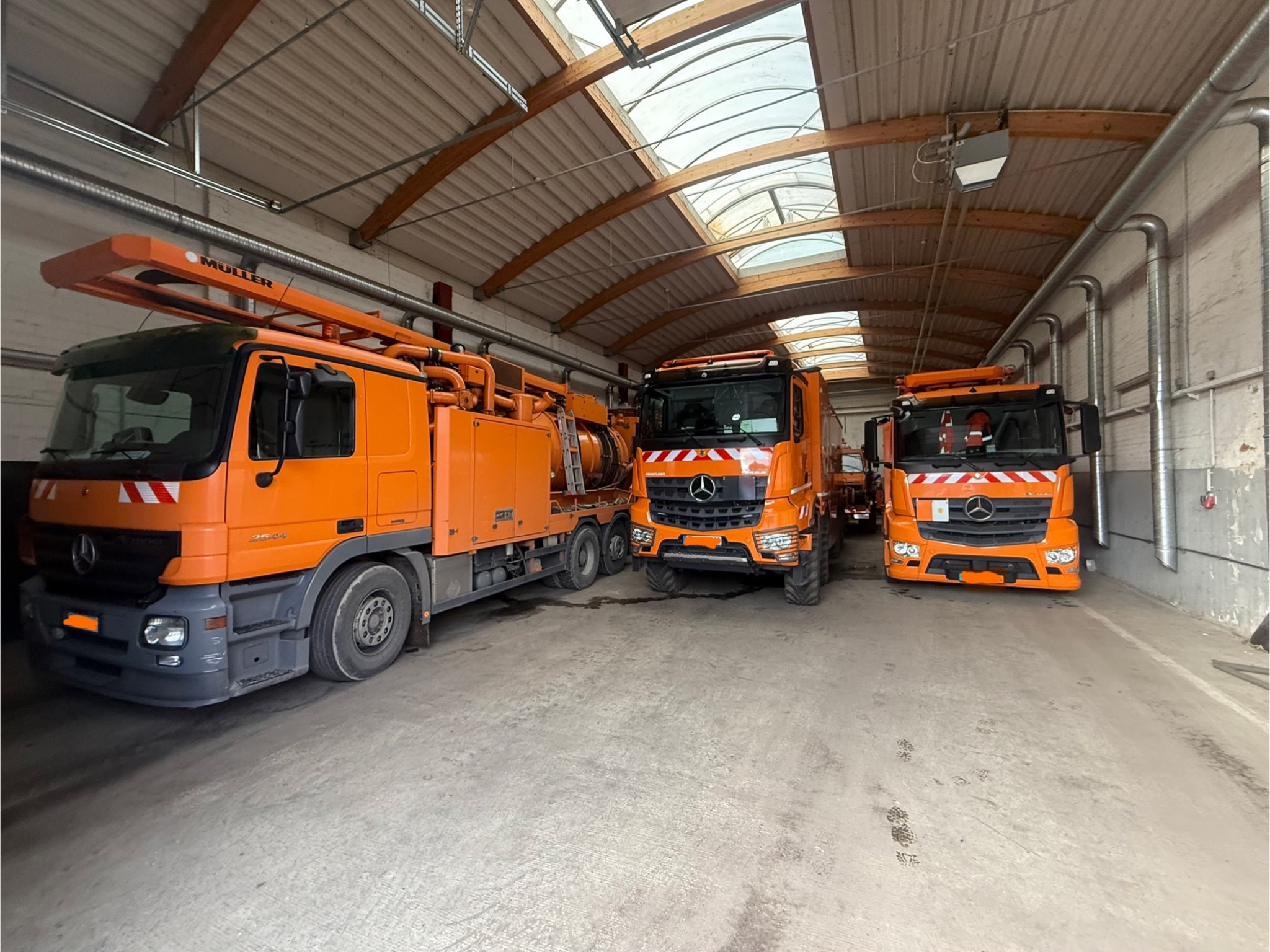 Pomarańczowe pojazdy specjalistyczne marki Mercedes-Benz w hali z drewnianą konstrukcją dachu i świetlikami. Jeden z pojazdów ma zamontowane rury i zbiornik.