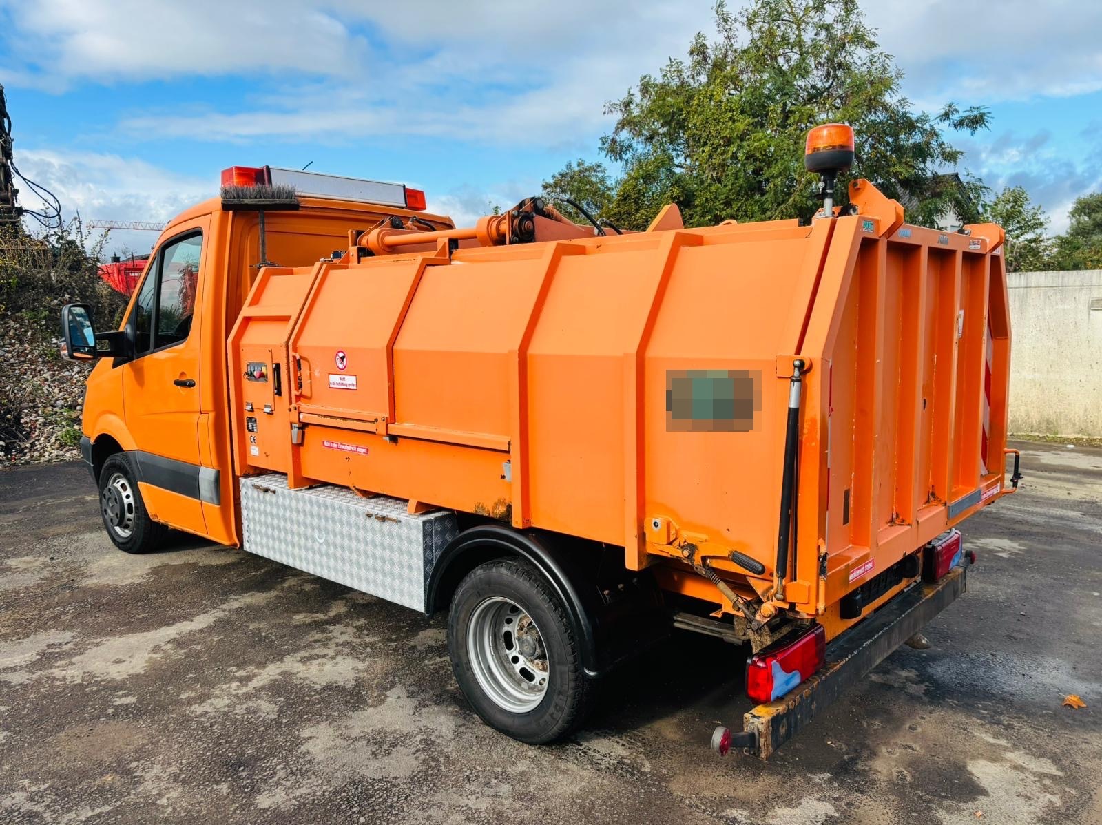 Pomarańczowa ciężarówka do czyszczenia ulic z zamontowaną szczotką i zbiornikiem, zaparkowana na asfalcie, widok z boku, w tle zieleń i błękitne niebo.