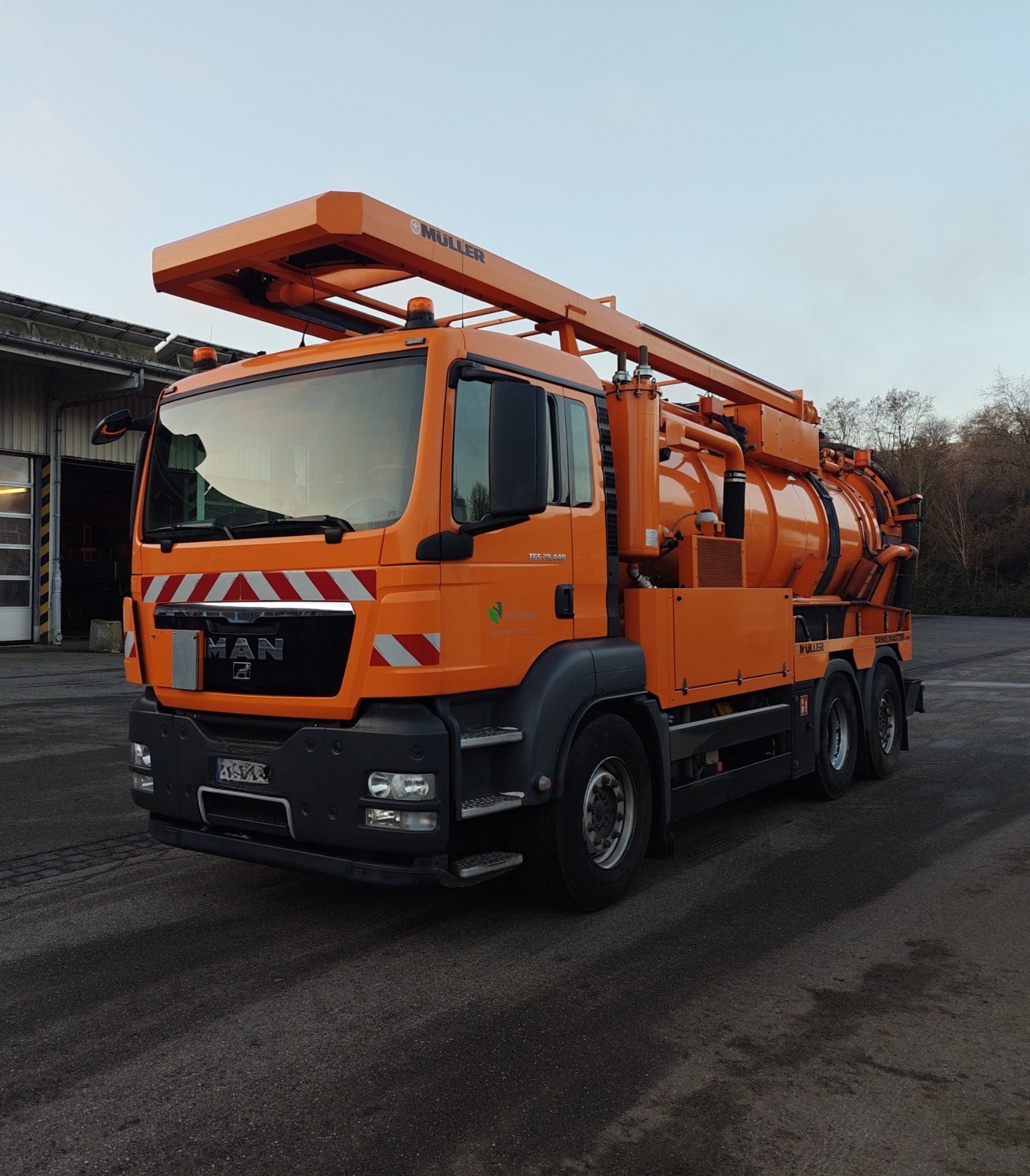 Pomarańczowy samochód asenizacyjny MAN TGS 28.440 z nadbudową Muller Canalmaster, zaparkowany na placu, z widocznymi elementami roboczymi i logo producenta.