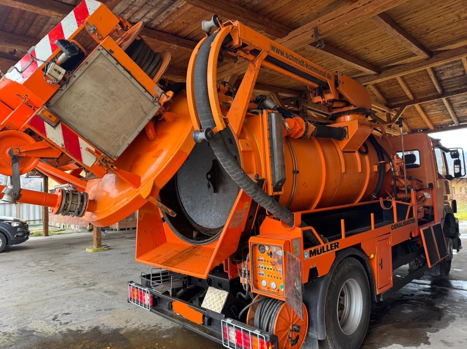 Pomarańczowy pojazd specjalistyczny z otwartym zbiornikiem pod drewnianą wiatą, prawdopodobnie do czyszczenia kanalizacji, z widocznymi elementami sterującymi i wężami.