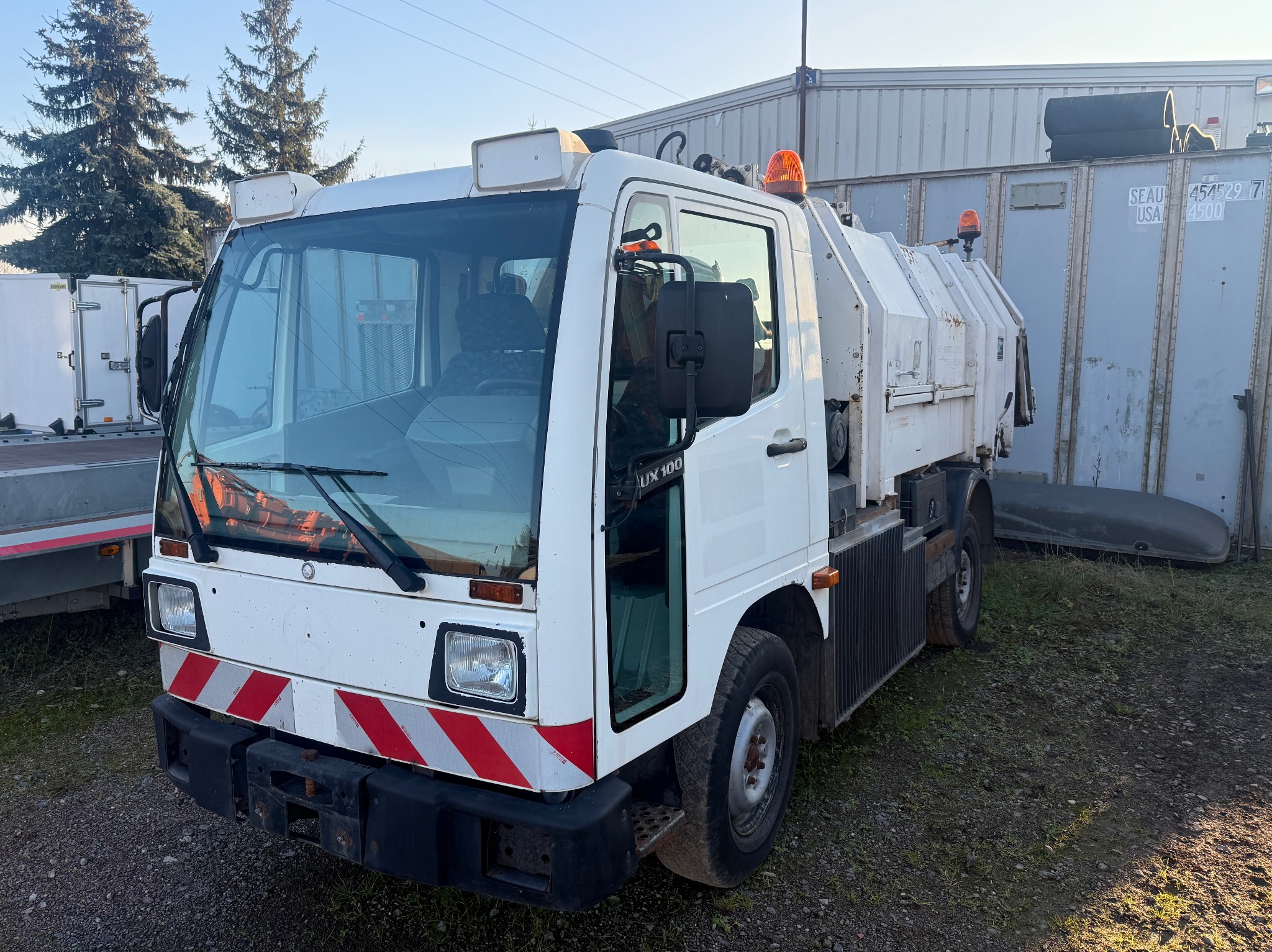 Biała zamiatarka uliczna LUX 100 z otwartymi drzwiami, zaparkowana na żwirowym podłożu, z widocznymi elementami ostrzegawczymi i kontenerami w tle.