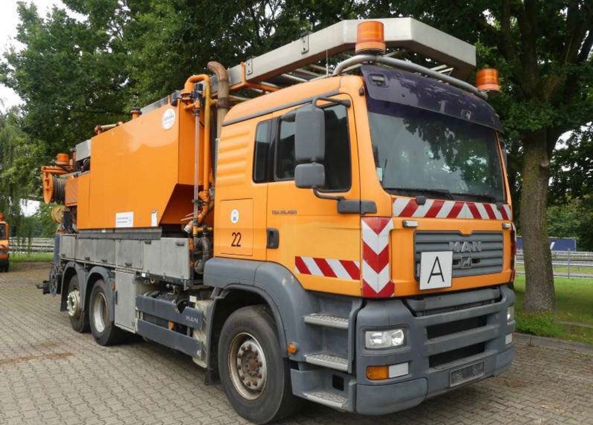 Pomarańczowy samochód ciężarowy MAN z urządzeniem do czyszczenia kanalizacji, zaparkowany na kostce brukowej, z zielonymi drzewami w tle. Widoczne oznaczenia ostrzegawcze.