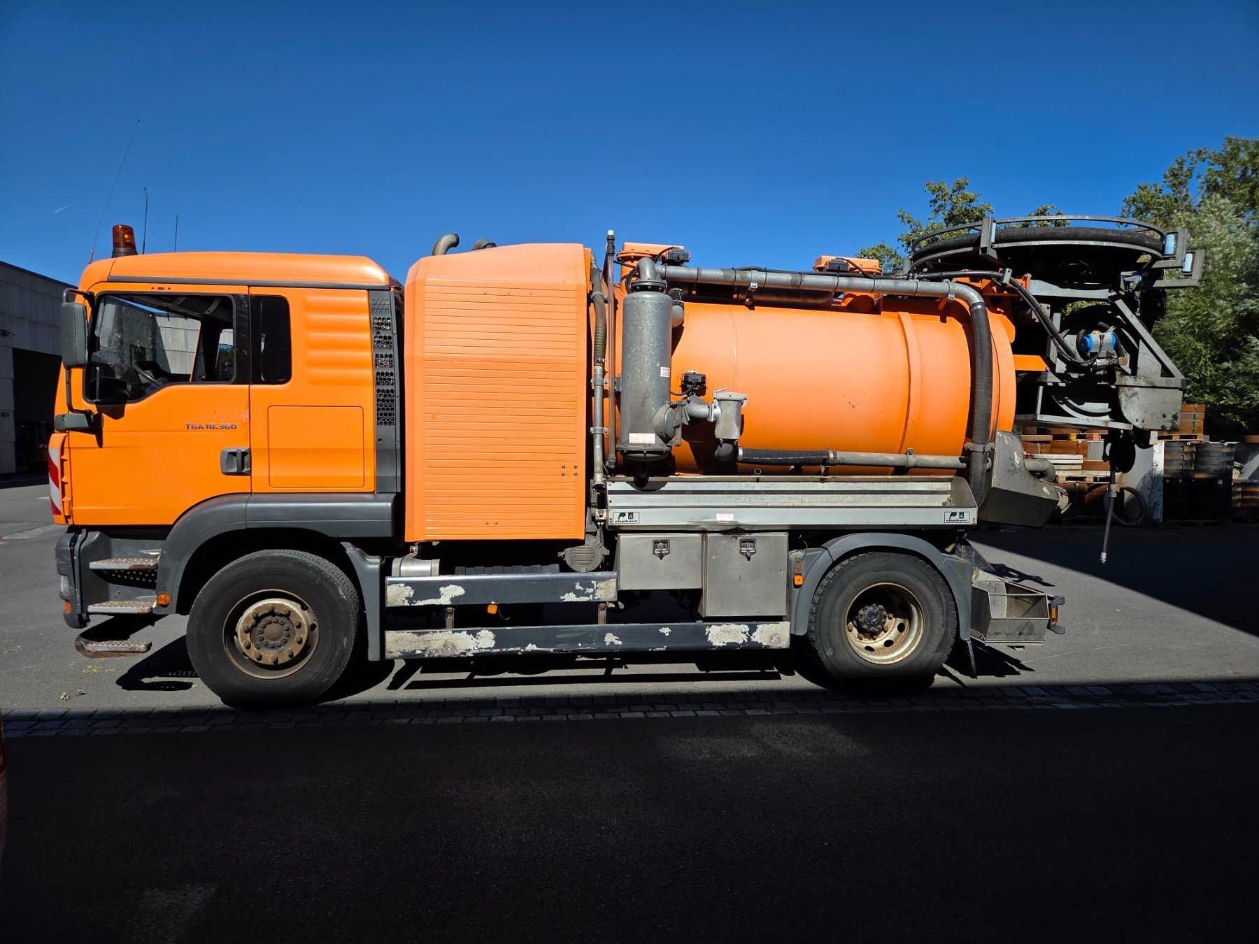 Pomarańczowy samochód asenizacyjny MAN TGA 18.360 z dużym zbiornikiem i elementami instalacji, zaparkowany na asfalcie w słoneczny dzień.