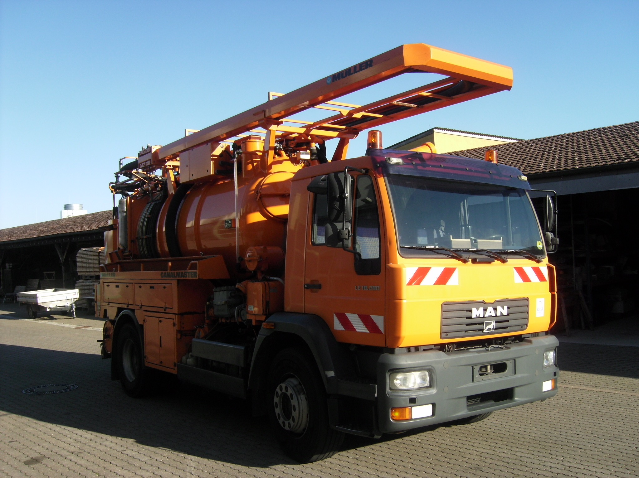 Pomarańczowy samochód asenizacyjny MAN z nadbudową Canalmaster, zaparkowany na brukowanej powierzchni w słoneczny dzień. Widoczny fragment budynku w tle.