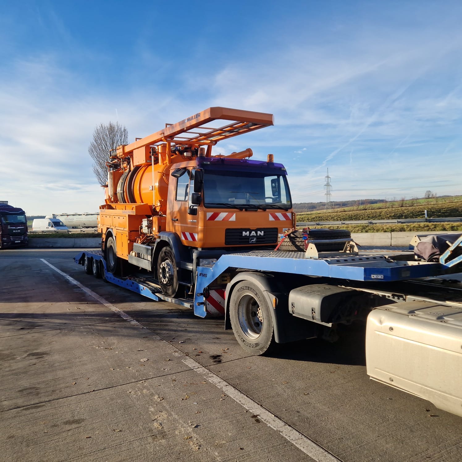 Pomarańczowy samochód ciężarowy MAN z zabudową do oczyszczania kanalizacji na niskopodwoziowej platformie transportowej, zaparkowany na betonowym placu.