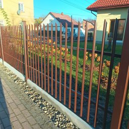 Ogrodzenia Gielarowski Tomasz - Nowoczesne ogrodzenie palisadowe w kolorze brązowym, z widocznymi elementami montażowymi, na tle zielonego trawnika i zabudowy jednorodzinnej w słoneczny dzień.