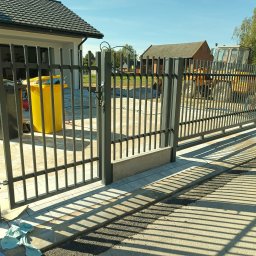 Ogrodzenia Gielarowski Tomasz - Nowoczesna, metalowa brama wjazdowa z pionowymi szczeblami, w tle dom z ciemną dachówką i żółty traktor. Cienie bramy na podjeździe.
