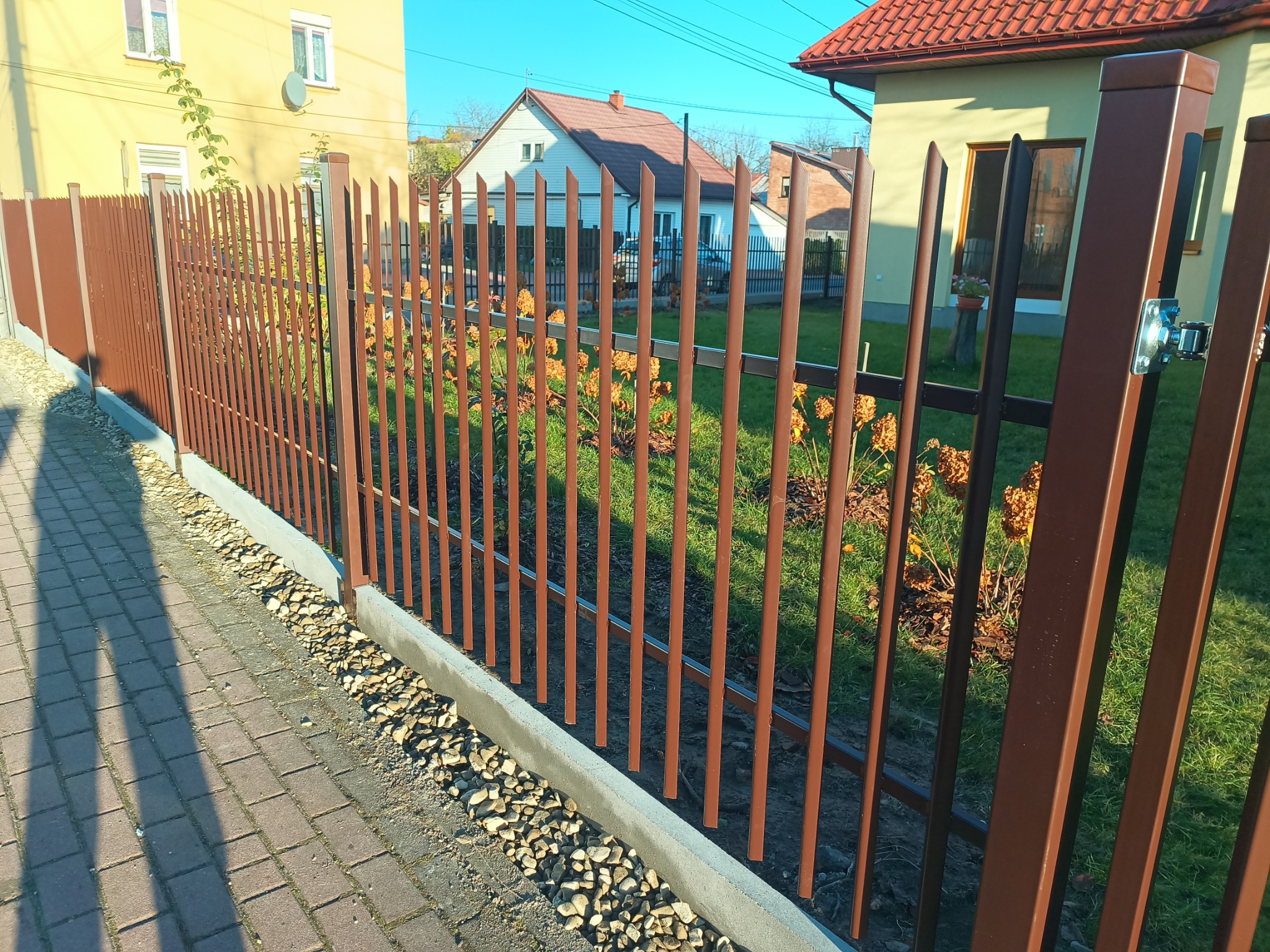 Nowoczesne ogrodzenie palisadowe w kolorze brązowym, z widocznymi elementami montażowymi, na tle zielonego trawnika i zabudowy jednorodzinnej w słoneczny dzień.