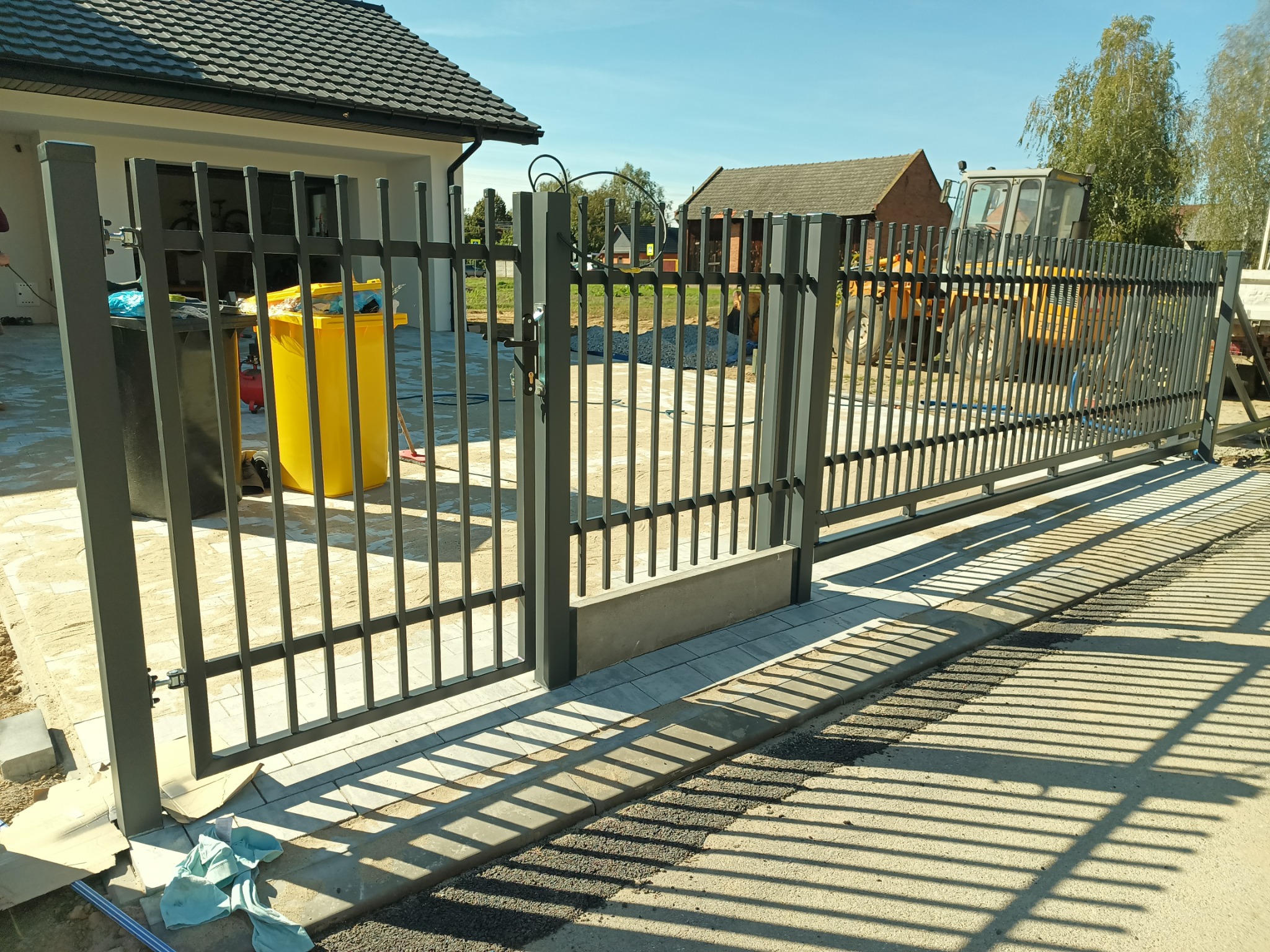 Nowoczesna, metalowa brama wjazdowa z pionowymi szczeblami, w tle dom z ciemną dachówką i żółty traktor. Cienie bramy na podjeździe.