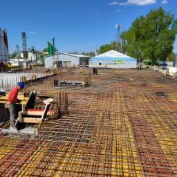 Praca na budowie: Zbrojarz w czerwonej bluzie i niebieskim kasku układa siatkę z prętów zbrojeniowych na żółtym szalunku, w tle budynki przemysłowe i błękitne niebo.