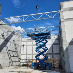 Montaż stalowej konstrukcji dachu hali z użyciem podnośnika nożycowego Mateco na tle muru z betonu komórkowego i błękitnego nieba.