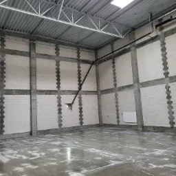 Wnętrze surowego, murowanego budynku w trakcie budowy z widocznymi ścianami z białych bloczków i betonowymi wzmocnieniami, mokrą betonową posadzką, metalową konstrukcją dachu i oświetleniem górnym.