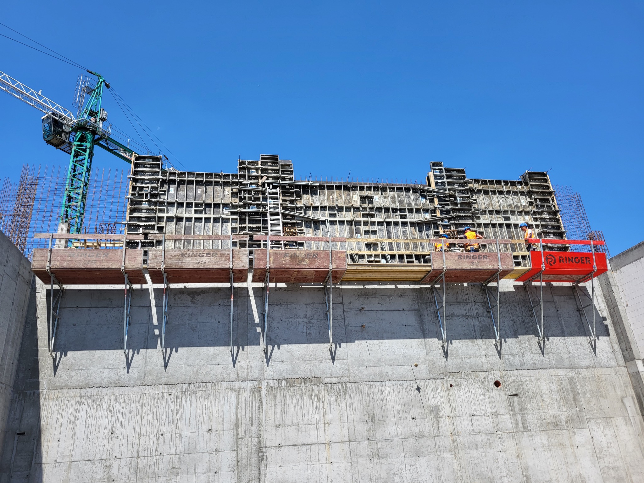 Wysoki betonowy mur z zamontowanym szalunkiem i rusztowaniami marki Ringer, na górze pracują robotnicy w pomarańczowych kaskach, w tle widoczny dźwig budowlany i błękitne niebo.
