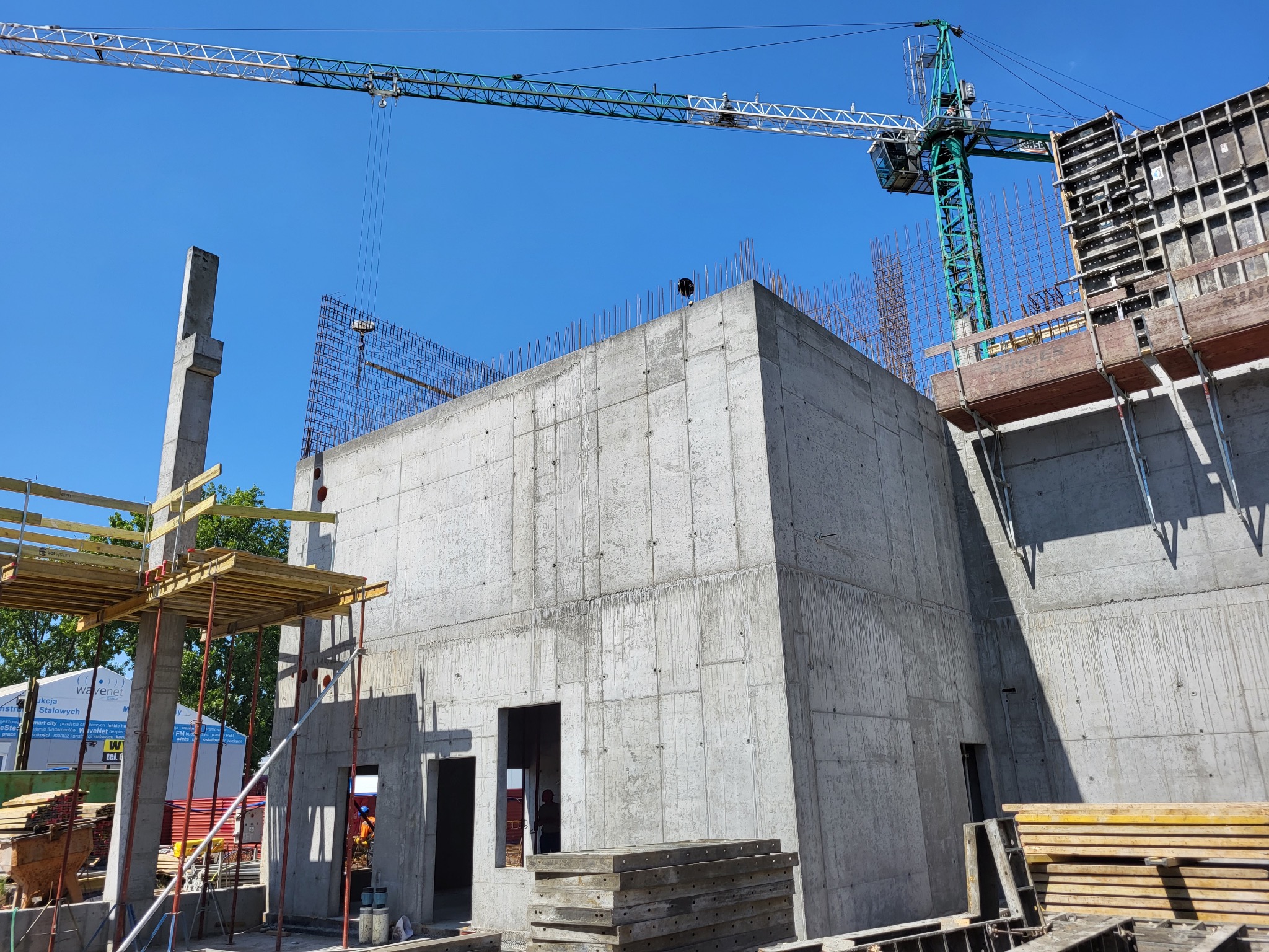 Surowy budynek żelbetowy w trakcie budowy, widoczny dźwig budowlany, szalunki, zbrojenia i betonowy słup na tle błękitnego nieba.