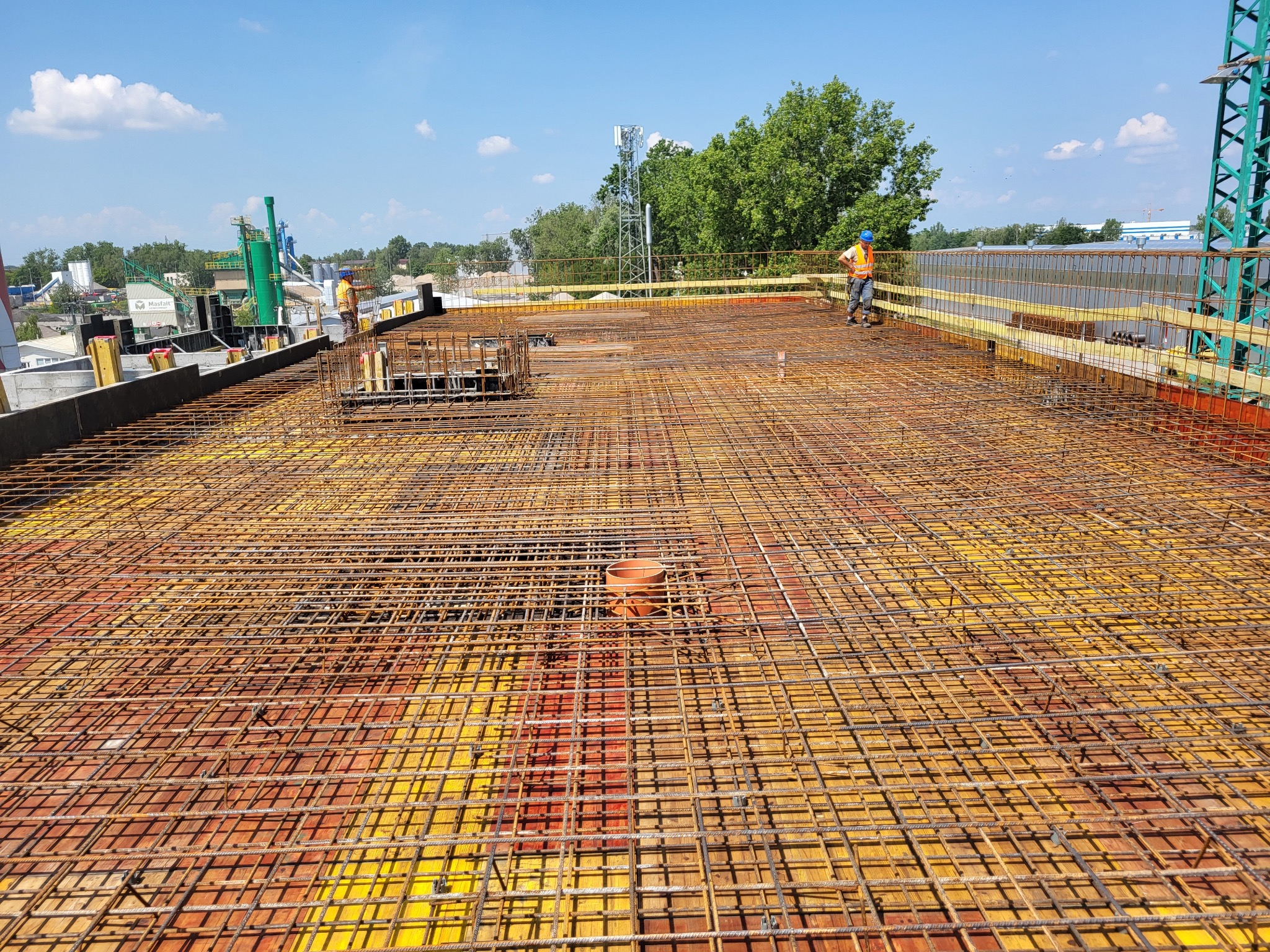 Siatka zbrojeniowa na żółtym szalunku, z pracownikiem w oddali, przygotowana do zalania betonem na budowie w słoneczny dzień.
