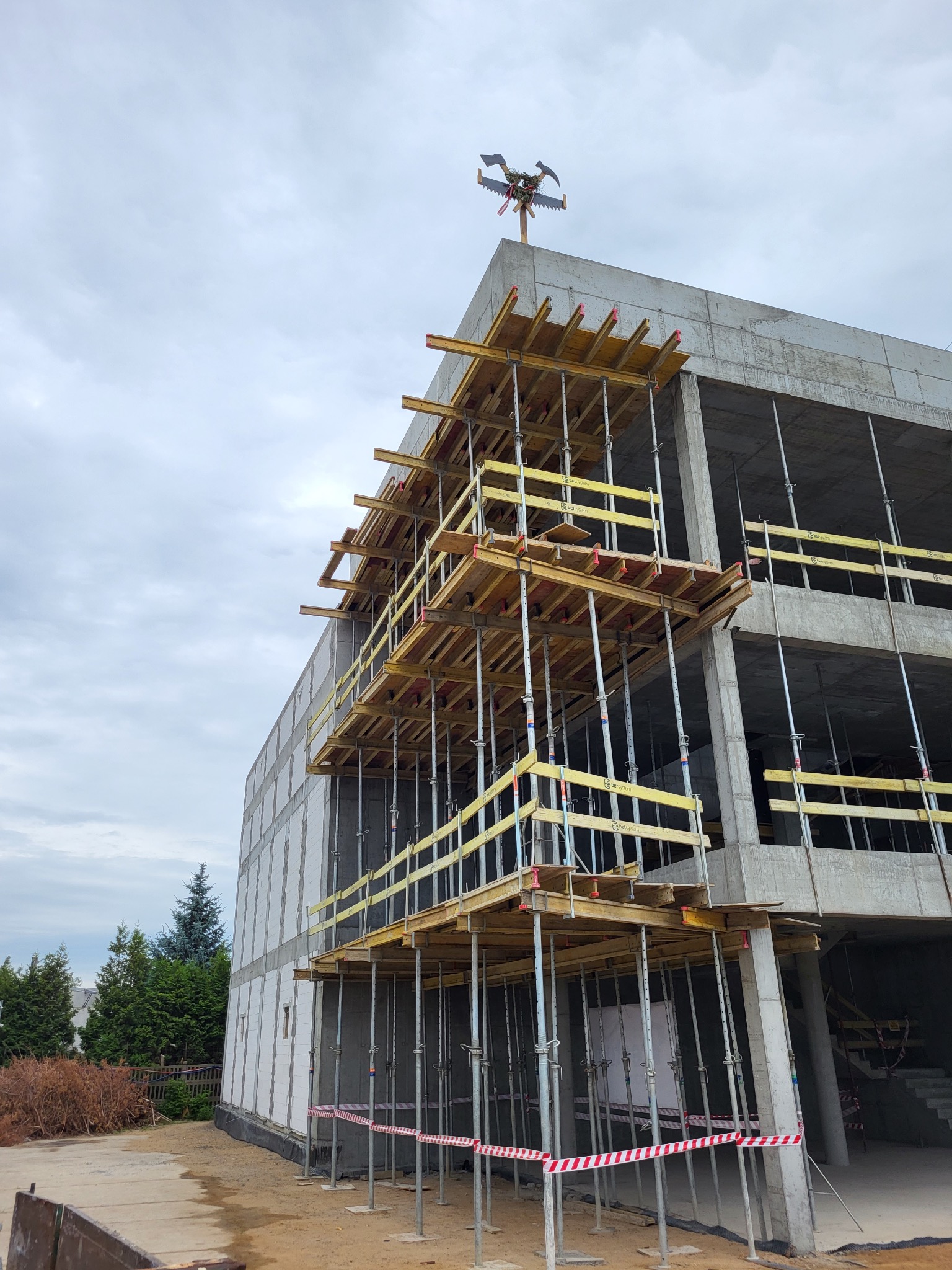 Surowy budynek żelbetowy w trakcie budowy, z widocznym szalunkiem i podporami, na szczycie budynku wieniec z narzędzi. Tło stanowi pochmurne niebo.